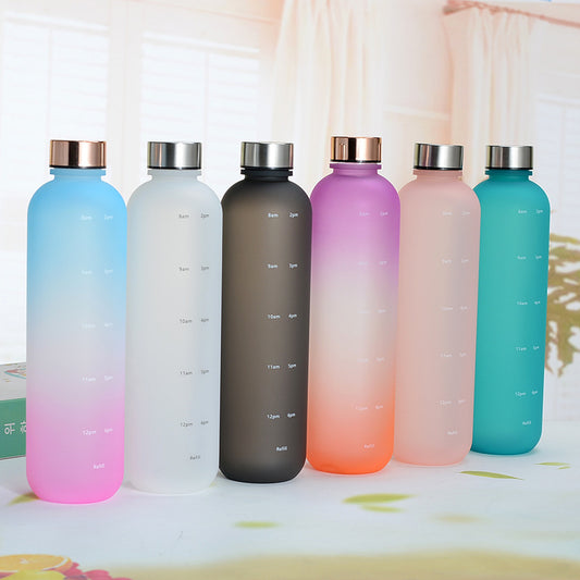Eine Reihe von sechs schlanken, farbverlaufenden Trinkflaschen mit Zeitmarkierungen. Die Flaschen haben verschiedene Farbverlaeufe: blau zu rosa, grau, dunkelgrau, lila zu rosa, rosa und blau-gruen. Sie stehen auf einem Tisch mit einer hellen, unscharfen Umgebung im Hintergrund.