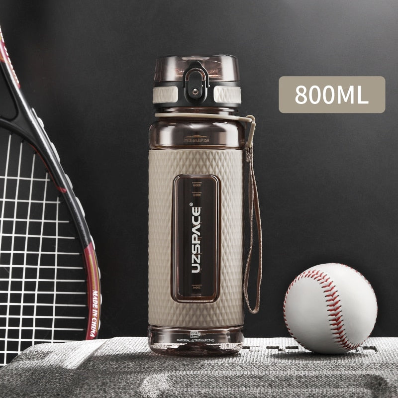 Eine beige Sport-Trinkflasche mit einem Fassungsvermoegen von 0.8 Litern, gekennzeichnet durch das Logo "UZSPACE" im klaren Sichtfenster, platziert zwischen einem Tennisracket und einem Baseball auf einer grauen strukturierten Oberflaeche vor einem schwarzen Hintergrund.