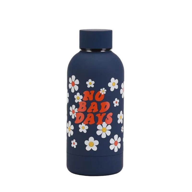 Eine dunkelblaue Kinder-Thermosflasche aus Edelstahl mit dem Aufdruck "NO BAD DAYS" umgeben von weißen und orangefarbenen Blumen.