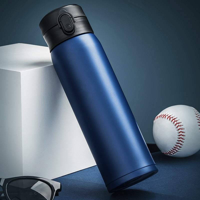 Eine blaue Edelstahl-Thermosflasche mit einem schwarzen Deckel, platziert neben einem Baseball und einer Sonnenbrille auf einem dunklen Untergrund vor einem hellen, geometrischen Hintergrund.