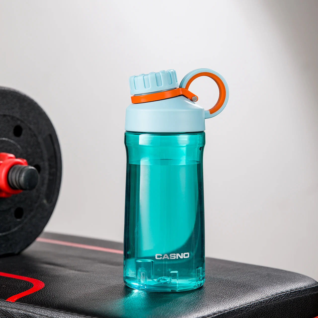 Eine türkis, transparente Sporttrinkflasche auf einer Fitnessbank. Die Flasche hat einen schwarzen Schraubverschluss mit einem Henkel und ist mit dem Wort der Marke "CASNO" beschriftet. Im Hintergrund ist eine Hantel mit roten Details zu sehen.
