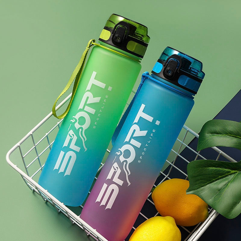 Zwei Sporttrinkflaschen mit Farbverlauf, eine in Gruen und die andere in Blau, in einem Einkaufskorb neben frischen Zitronen und einem grünen Blatt, vor einem olivgruenen Hintergrund.