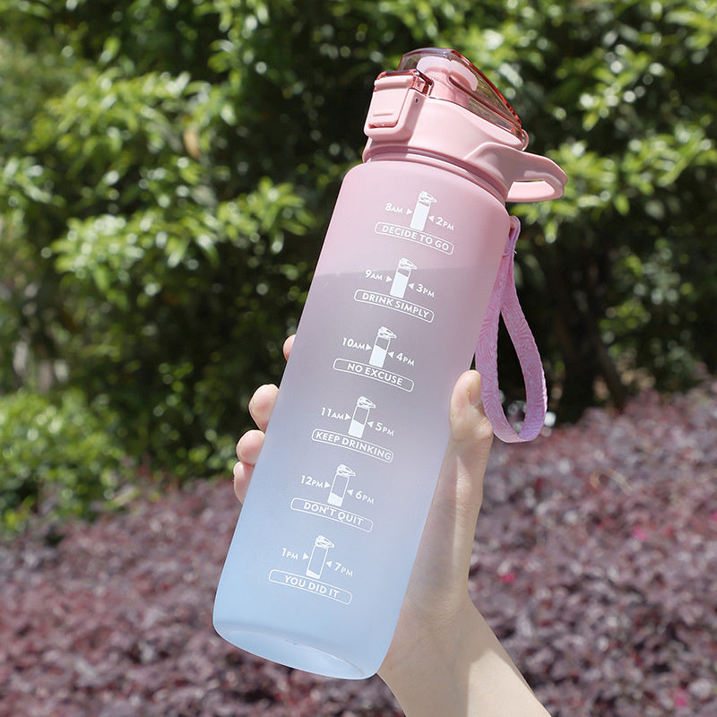 Eine Hand haelt eine rosa Trinkflasche mit einem Farbverlauf nach Blau und motivierenden Zeitmarkierungen vor einem gruenen und rotlaubigen Hintergrund.