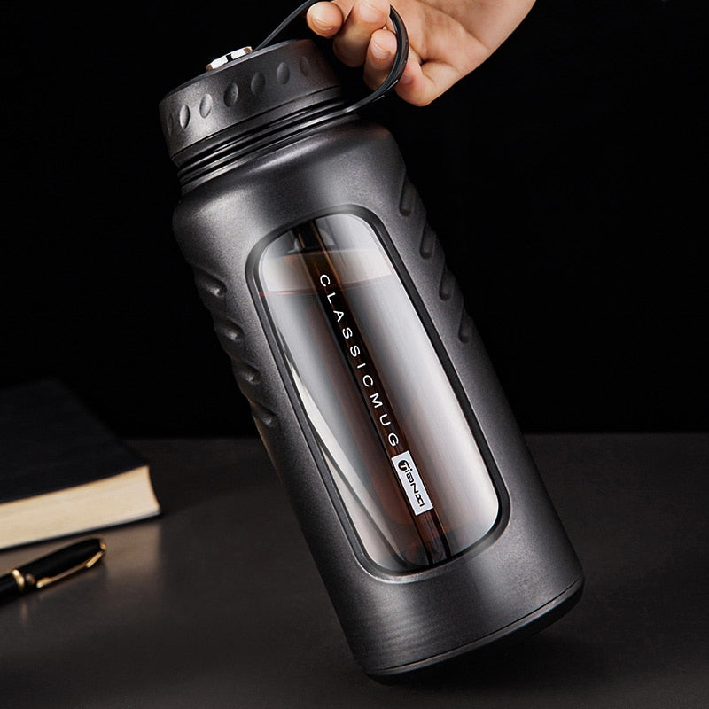 Schwarze, doppelwandige Glas-Trinkflasche mit Tragegriff und integriertem Teesieb, platziert auf einem dunklen Untergrund neben einem stilvollen Stift und Notizbuch.