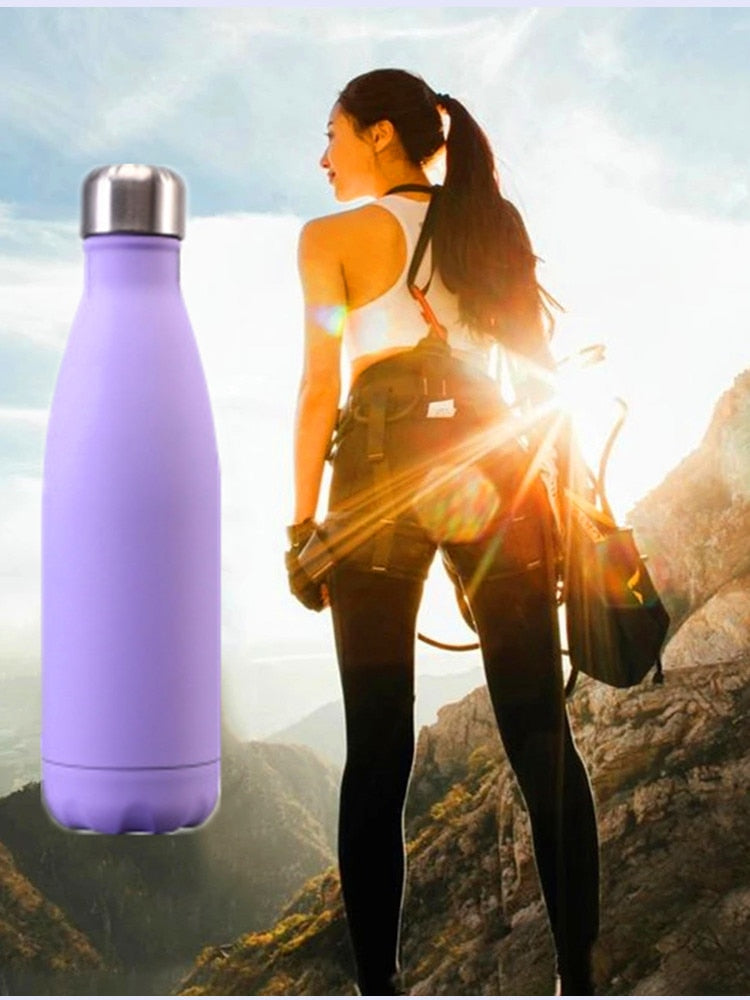 Eine lila Edelstahl-Thermosflasche im Vordergrund und im Hintergrund eine Person, die mit einem Rucksack in einer bergigen Landschaft wandert.