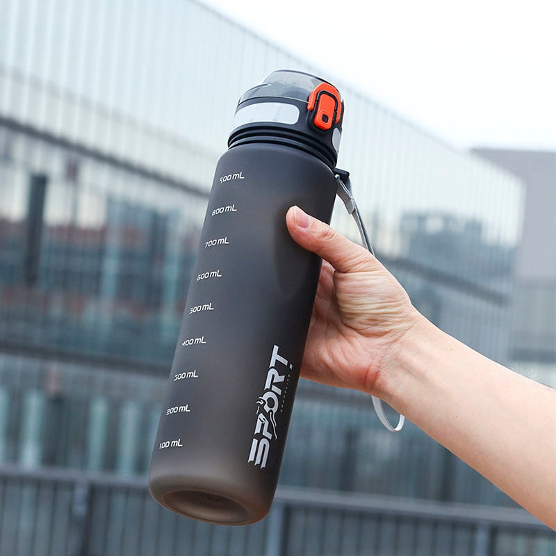 Eine Hand haelt eine schwarze Sporttrinkflasche mit Volumenmarkierungen und dem Aufdruck 'SPORT', vor einem unscharfen staedtischen Hintergrund.