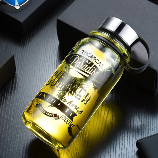 Transparente Trinkflasche aus Glas mit sommerlichem Aufdruck "TROPICAL SURF PARADISE" und "SUMMER is here ENJOY IT" in Gelb, platziert neben einer schwarzen Box auf einem dunkelgrauen Untergrund.