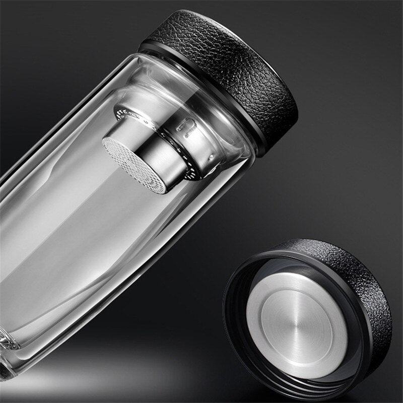 Eine transparente, laengliche Glasflasche mit einem integrierten Teesieb und einem schwarzen, strukturierten Deckel, beides hervorgehoben auf einem dunklen Hintergrund.