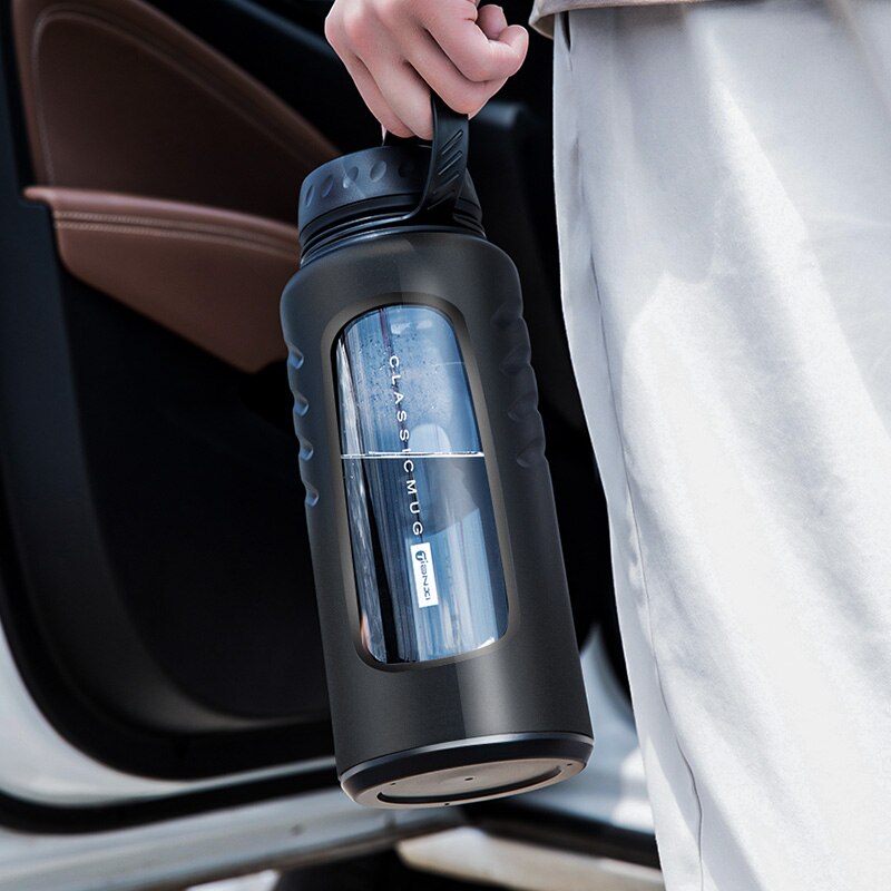 Stilvolle, schwarze, doppelwandige Glasflasche mit Teesieb und praktischem Tragegriff, gehalten von einer Person neben einem weissen Auto.