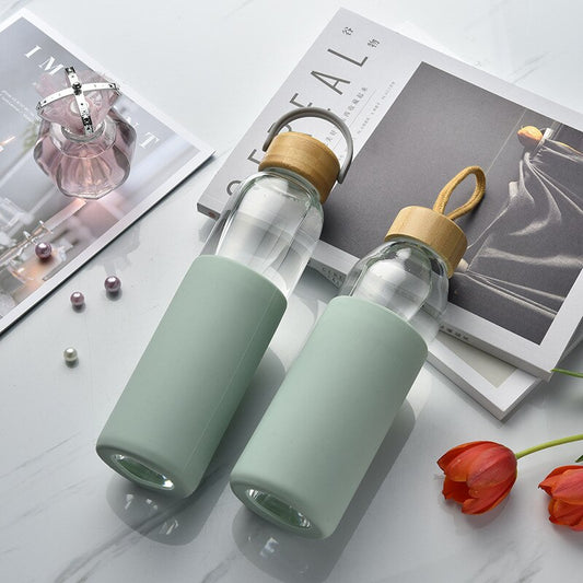 Zwei trendige Glasflaschen mit Silikonschutzhuelle und Bambusdeckel auf einem Tisch, neben einer Modezeitschrift und roten Tulpen.