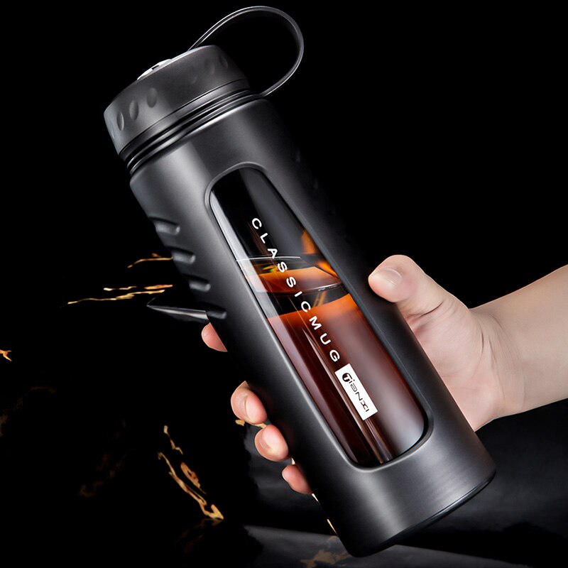 Hand haelt eine Moderne, schwarze, doppelwandige Trinkflasche aus Glas mit integriertem Teesieb, Griff und abgebildet neben einer stilvollen Uhr und einer Tasse Tee.