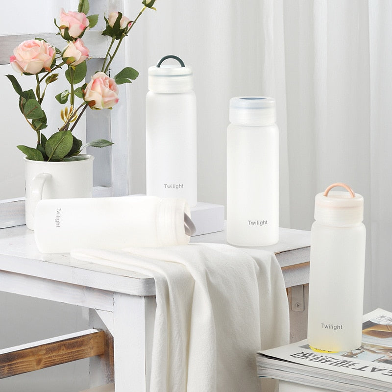 Vier weiße, matte Glasflaschen mit der Aufschrift 'Twilight', zwei mit Deckeln in verschiedenen Farben, auf einem weißen Tisch neben einem Topf mit rosa Rosen und einem weißen Tuch.