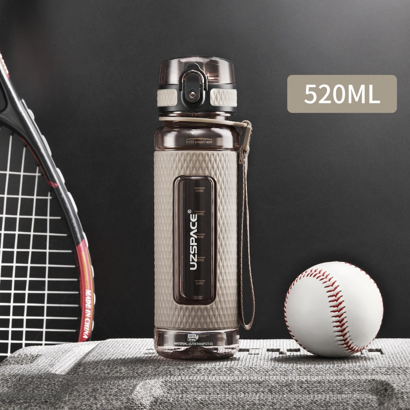 Eine beige Sport-Trinkflasche mit einem Fassungsvermoegen von 0.52 Litern, gekennzeichnet durch das Logo "UZSPACE" im klaren Sichtfenster, platziert zwischen einem Tennisracket und einem Baseball auf einer grauen strukturierten Oberflaeche vor einem schwarzen Hintergrund.