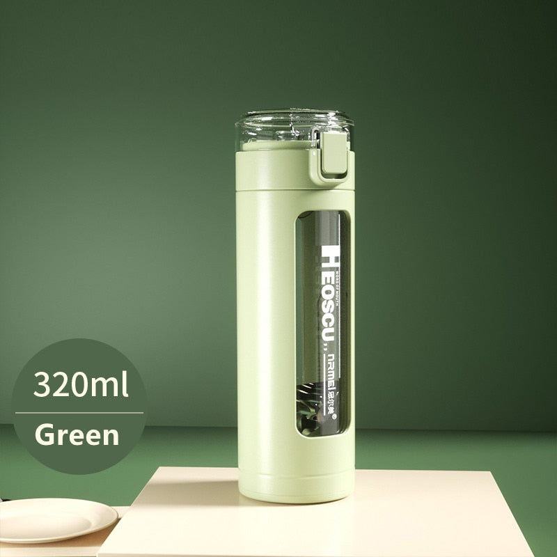 Eine gruene Glastrinkflasche mit einem Fassungsvermoegen von 320 ml, ausgestattet mit einem aufklappbaren Deckel und einem eingebauten Strohhalm. Die Flasche steht auf einer cremefarbenen Oberflaeche vor einem einfarbigen gruenen Hintergrund.