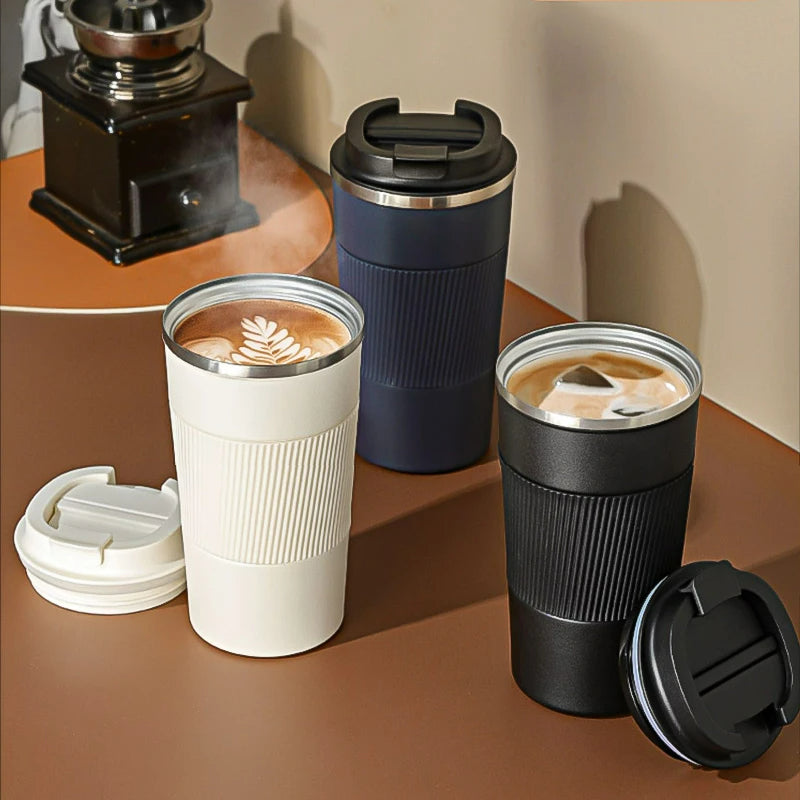 Drei Thermobecher fuer Kaffee in Weiß, Blau und Schwarz. Die weiße und schwarze Tasse sind geoeffnet und zeigen kunstvoll dekorierte Kaffeegetraenke, während die blaue Tasse einen verschlossenen Deckel mit Trinkoeffnung hat.