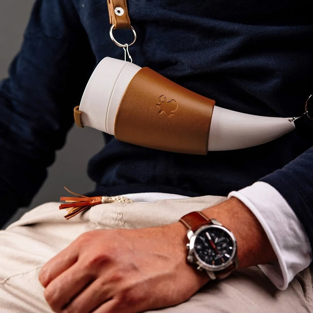 Einen hornfoermigen Kaffee- oder Thermosbecher mit einem braunen Ledergriff und weißem Koerper, der von einer Person gehalten wird, die eine Uhr traegt.