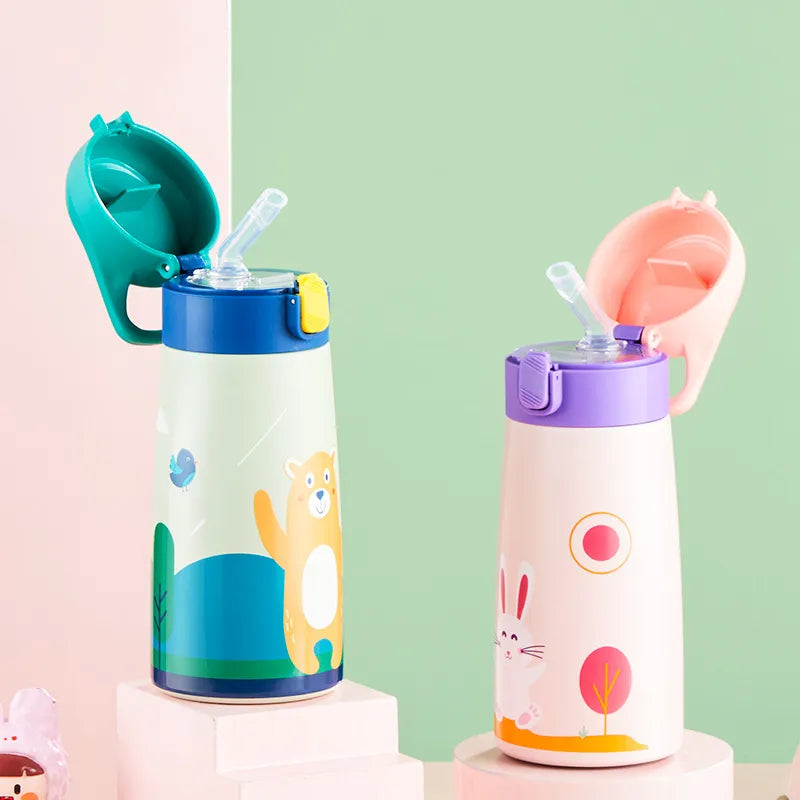Zwei Thermosflaschen fuer Kinder, die eine mit einem Baeren in einem blauen Waldmotiv und die andere mit einem Hasen in einem hellen Motiv mit Sonne und Baeumen. Beide Flaschen haben einen integrierten Strohhalm und stehen auf einem Podest.