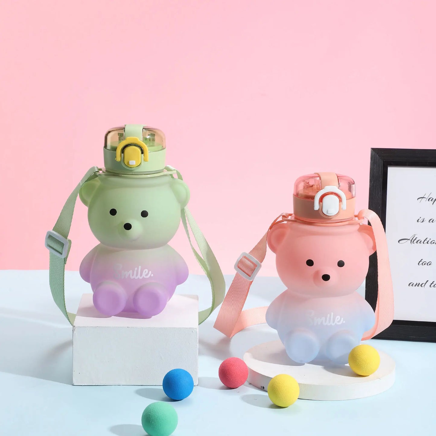 Zwei Teddybaer-Trinkflaschen, eine gruen und die andere rosa, mit der Aufschrift "Smile", stehen neben einander vor einem rosa Hintergrund. Beide haben Tragegurte und sind umgeben von bunten Baellen.