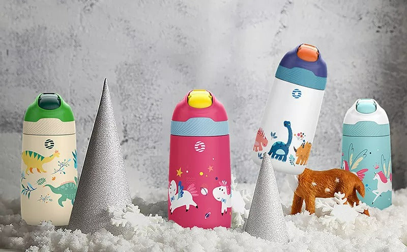Farbige Kinder-Thermosflaschen mit Tiermotiven, platziert zwischen dekorativen silbernen Tannenbaeumen auf kuenstlichem Schnee.