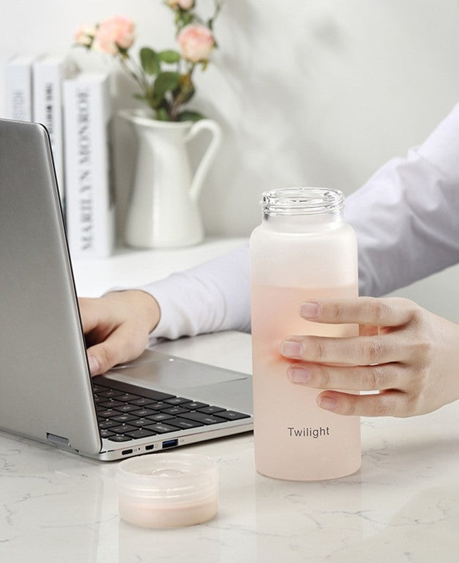 Person in einem weißen Hemd benutzt ein Laptop, haelt eine matte, rosa gefaerbte Glasflasche mit der Aufschrift 'Twilight', vor einem weißen Hintergrund mit Rosen und Buechern.