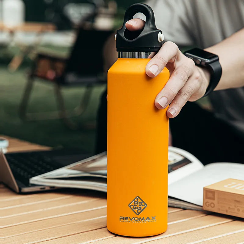Eine Hand haelt eine orange Edelstahl-Thermosflasche der Marke Revomax ueber einem Tisch, auf dem auch ein Laptop und ein geoeffnetes Buch liegen.