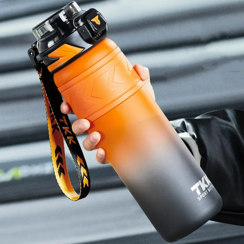 Orange-schwarze Sport-Trinkflasche mit 1 Liter Fassungsvermoegen und integriertem Strohhalm. Sie ist mit einem praktischen Tragegriff und einem orange-schwarzem Trageband ausgestattet. Die Flasche wird gehalten und ist vor einem unscharfen, grauen Hintergrund positioniert.