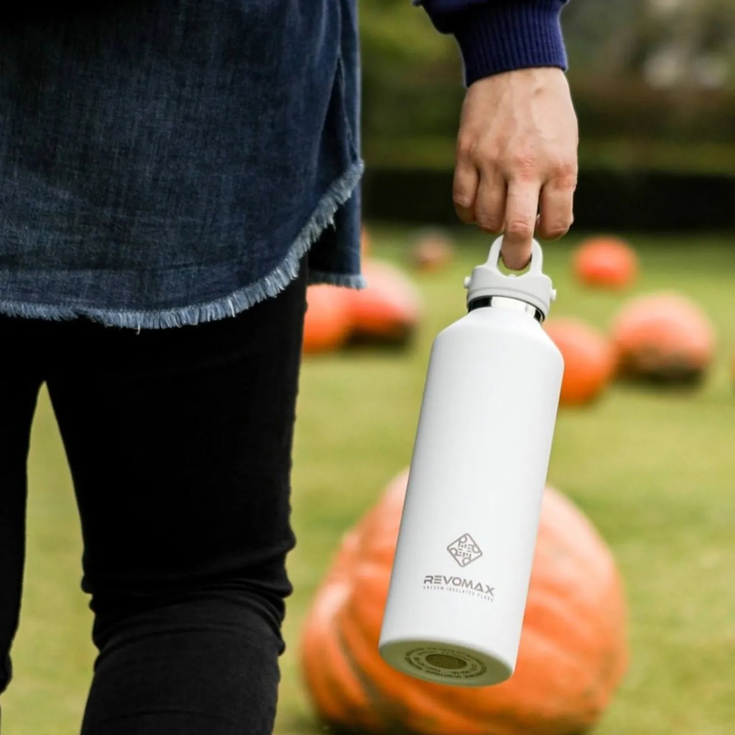 Eine Person haelt eine weiße Edelstahl-Thermosflasche der Marke Revomax, im Hintergrund sind Kuerbisse auf einem Rasen zu sehen.