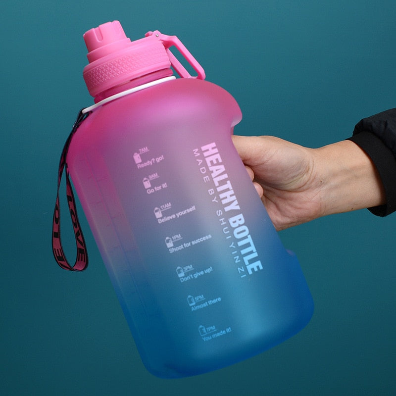 Eine Hand haelt eine grosse, pink blaue Sporttrinkflasche mit einem Henkel und aufgedrucktem Text "HEALTHY BOTTLE" und motivierenden Slogans zur Wasseraufnahme.