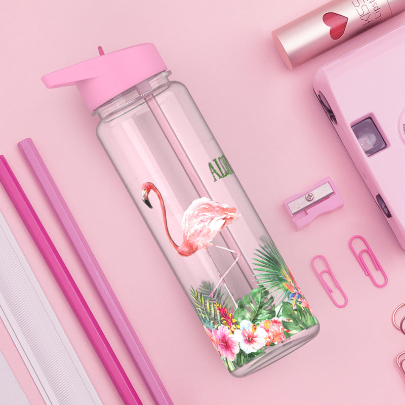 Eine Kindertrinkflasche mit einem rosafarbenen Deckel, auf der ein Flamingo und tropische Blumen abgebildet sind. Die Flasche ist Teil einer rosa getoenten Szene, die auch Schreibwaren wie Stifte, einen Spitzer, Bueroklammern, ein Notizbuch und einen Taschenrechner enthaelt.