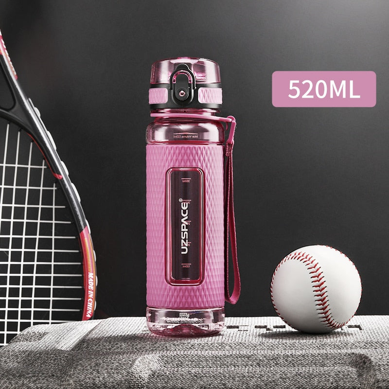 Eine pinke Sport-Trinkflasche mit einem Fassungsvermoegen von 0.52 Litern, gekennzeichnet durch das Logo "UZSPACE" im klaren Sichtfenster, platziert zwischen einem Tennisracket und einem Baseball auf einer grauen strukturierten Oberflaeche vor einem schwarzen Hintergrund.