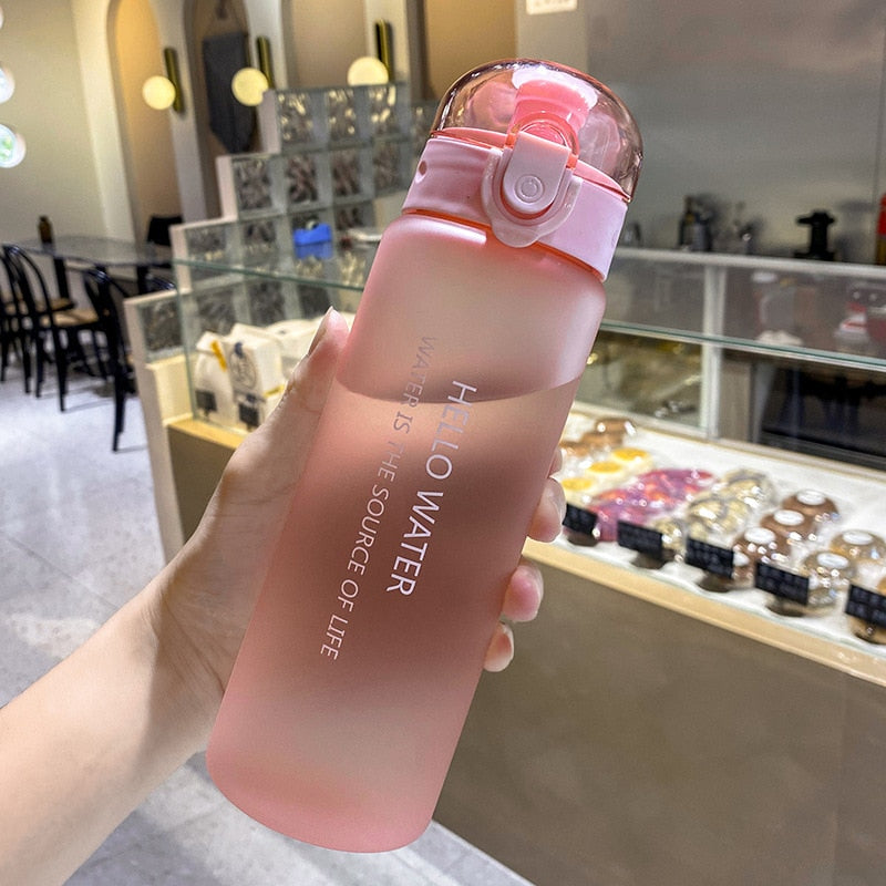 Eine Hand haelt eine pinke Sport-Trinkflasche mit einem Deckel und einem Druckknopfverschluss. Der Hintergrund ist unscharf, zeigt aber eine helle Inneneinrichtung, eines Cafés, mit Glasvitrinen und Leuchten.