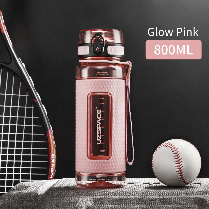 Eine pinke Sport-Trinkflasche mit einem Fassungsvermoegen von 0.8 Litern, gekennzeichnet durch das Logo "UZSPACE" im klaren Sichtfenster, platziert zwischen einem Tennisracket und einem Baseball auf einer grauen strukturierten Oberflaeche vor einem schwarzen Hintergrund.