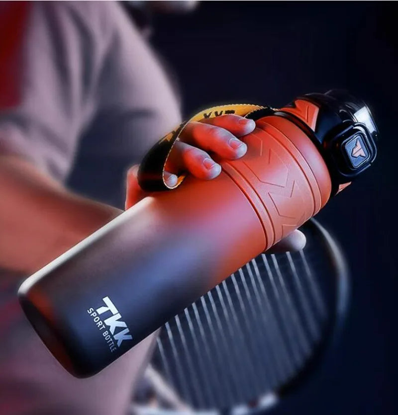 Hand haelt eine rot-schwarze Sport-Trinkflasche mit einem Fassungsvermoegen von 1 Liter. Die Flasche hat ein robustes Design mit einer strukturierten Oberflaeche fuer besseren Griff und eine auffaellige gelbe Schlaufe. Im Hintergrund ist schemenhaft ein Tennisschläger zu erkennen.