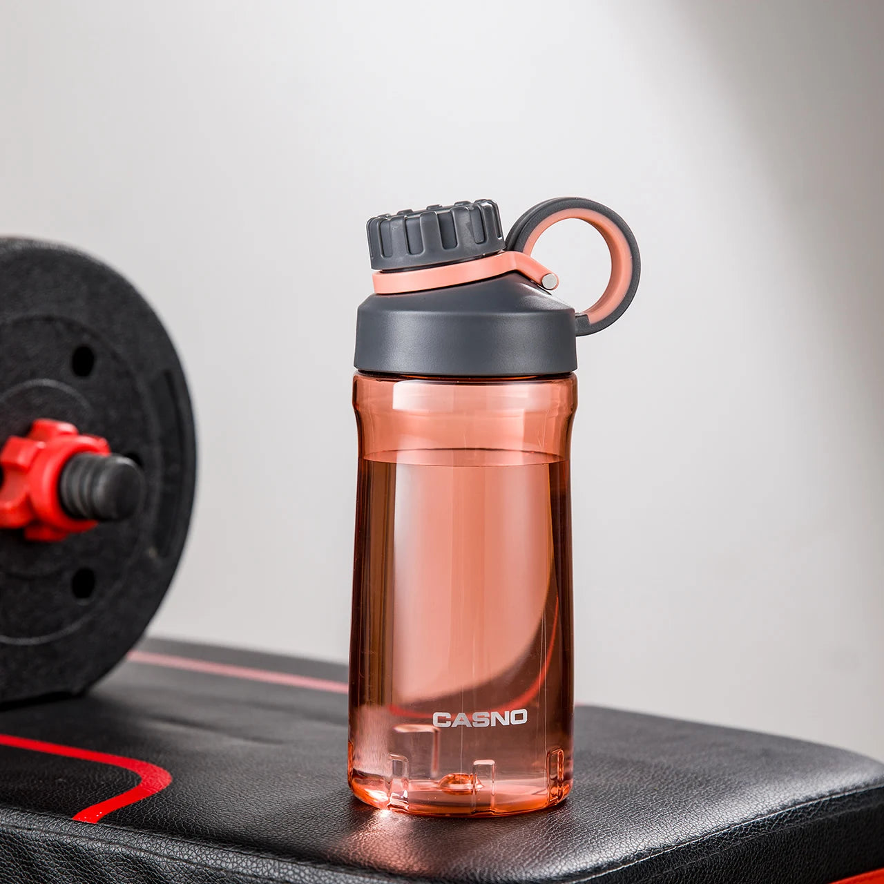 Eine rote, transparente Sporttrinkflasche auf einer Fitnessbank. Die Flasche hat einen schwarzen Schraubverschluss mit einem Henkel und ist mit dem Wort "SPORT" sowie der Marke "CASNO" beschriftet. Im Hintergrund ist eine Hantel mit roten Details zu sehen.