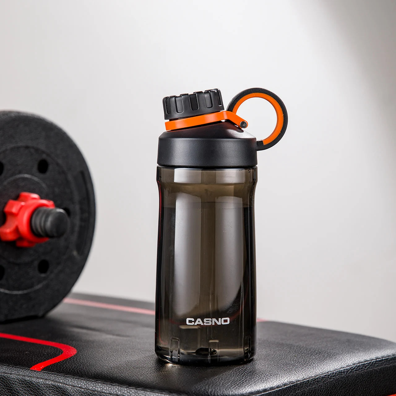Eine schwarze, transparente Sporttrinkflasche auf einer Fitnessbank. Die Flasche hat einen schwarzen Schraubverschluss mit einem Henkel und ist mit dem Wort "SPORT" sowie der Marke "CASNO" beschriftet. Im Hintergrund ist eine Hantel mit roten Details zu sehen.