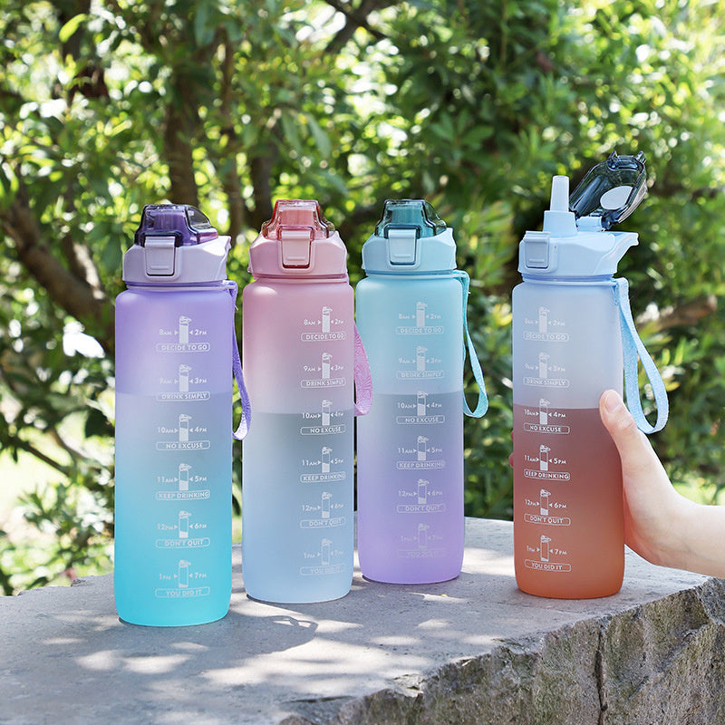 Vier farbverlaufende Sporttrinkflaschen mit Zeitmarkierungen, unterschiedlichen Deckelfarben und Tragegriffen, auf eine Stein vor einem gruenen, blaettrigen Hintergrund.
