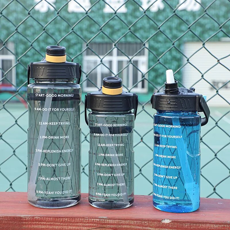 Drei Sporttrinkflaschen mit motivierenden Zeitmarkierungen, aufgestellt auf einer roten Oberflaeche vor einem Tennisplatzzaun.