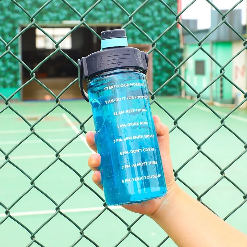 Eine Hand haelt eine blaue Sporttrinkflasche mit Zeitmarkierungen und motivierenden Spruechen vor einem Tennisplatzzaun.