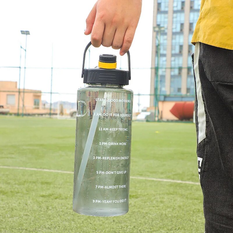 Eine Hand haelt eine klare Sporttrinkflasche mit gelbeschwarzem Deckel und Zeitmarkierungen, vor einem Fußballfeld mit Gebaeuden im Hintergrund.