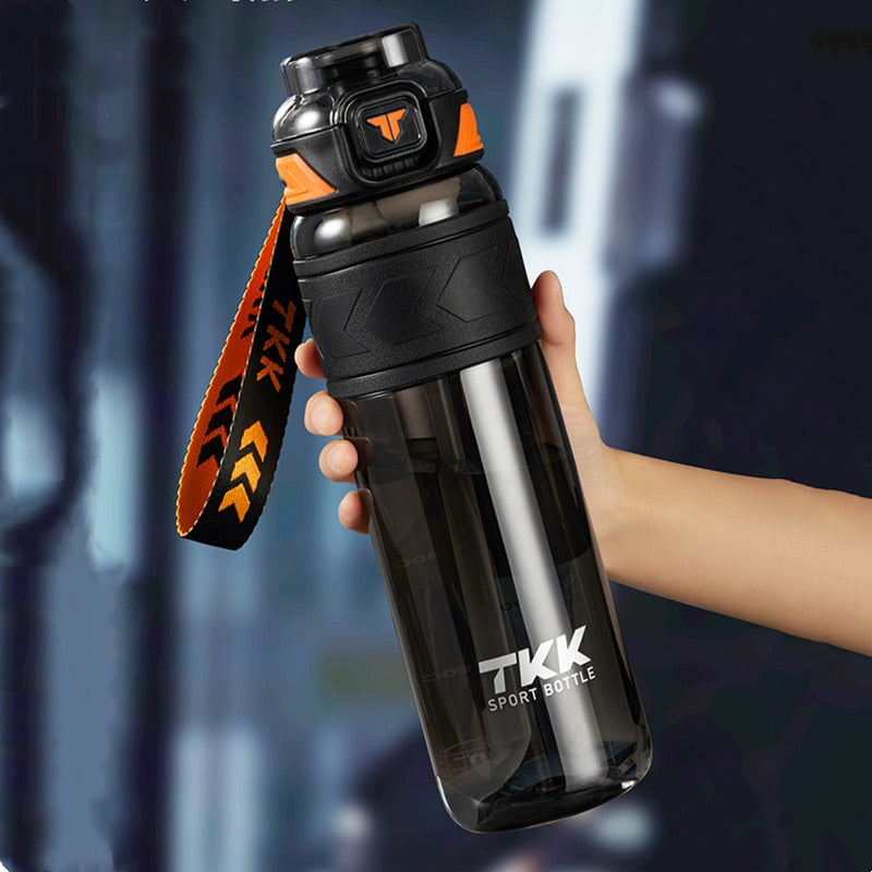 Eine Hand haelt eine transparente schwarze Sporttrinkflasche mit der Aufschrift TKK und einem offenen Deckel, aus dem eine Schlaufe ragt.