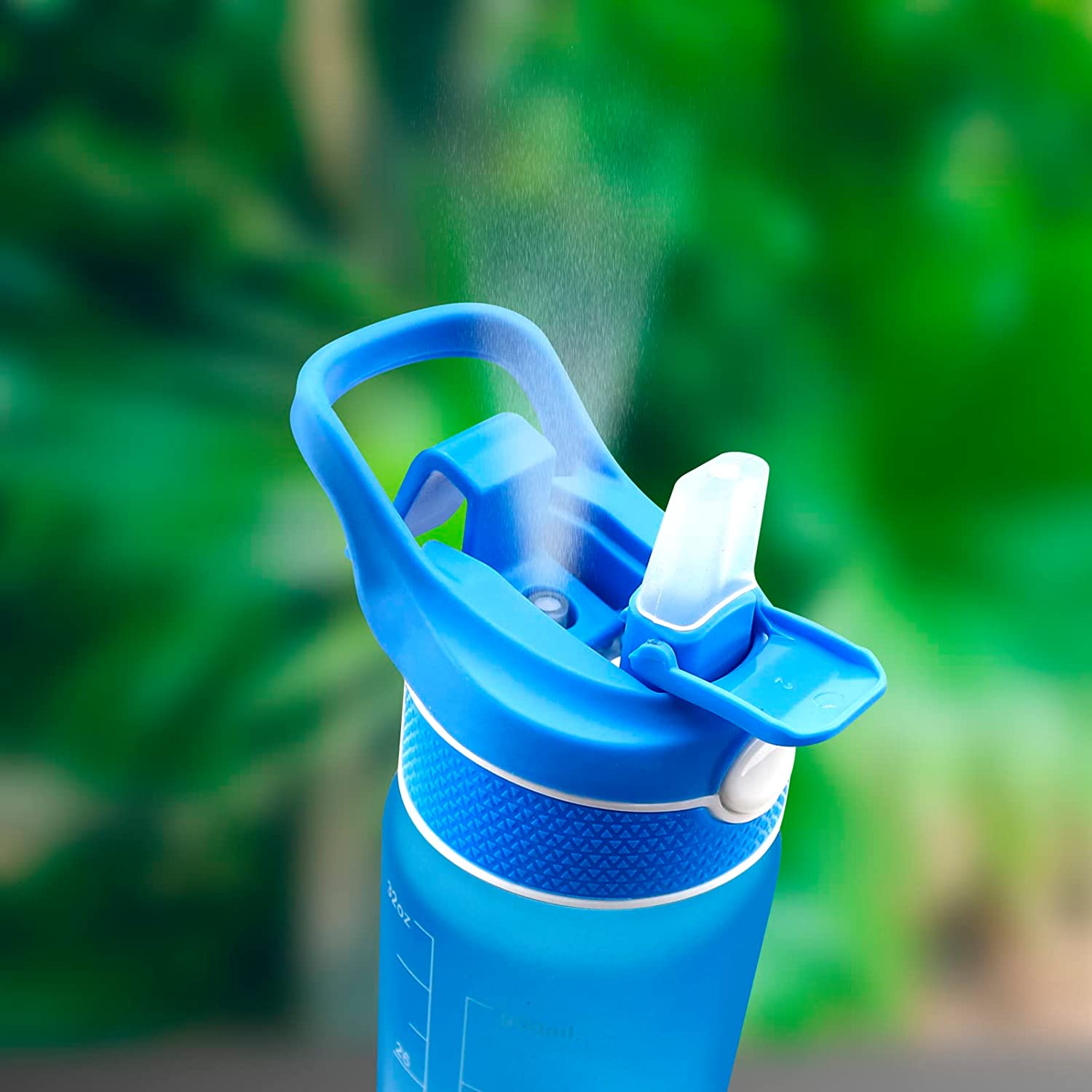 Eine blaue Sporttrinkflasche mit Spruehfunktion, aktiviert, um einen feinen Wassernebel zu verspruehen, vor einem unscharfen gruenen Hintergrund.