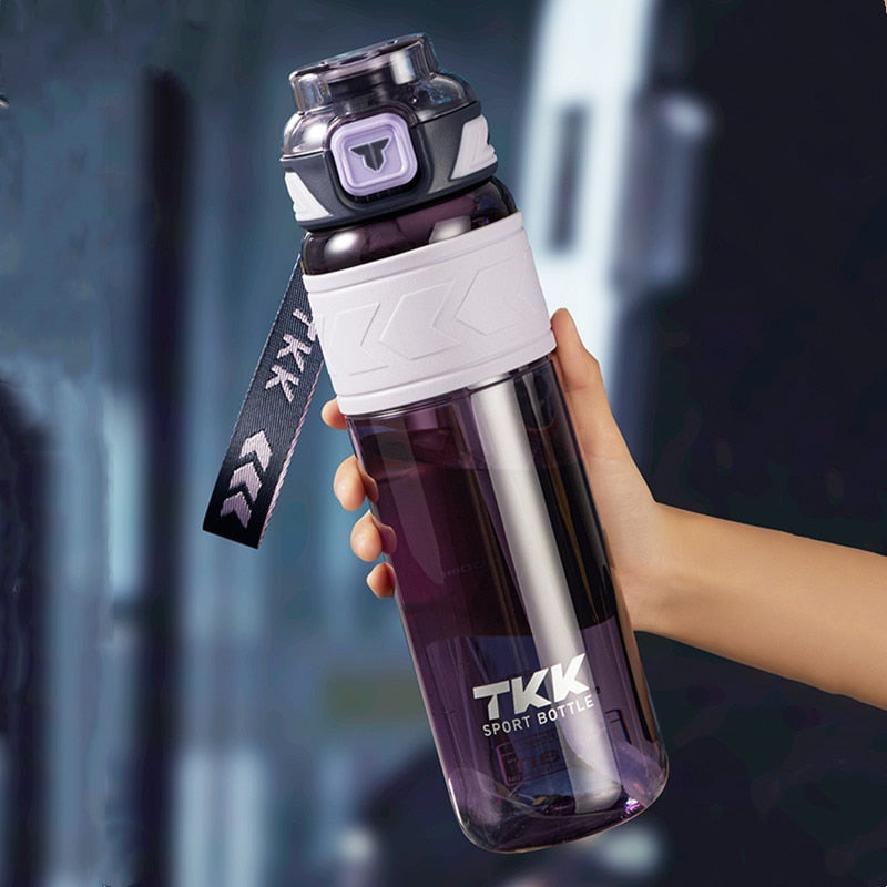 Eine Hand haelt eine transparente violette Sporttrinkflasche mit der Aufschrift TKK und einem offenen Deckel, aus dem eine Schlaufe ragt.