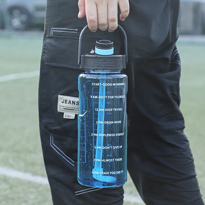 Eine Person mit schwarzer Hose haelt in der Hand eine blaue Sporttrinkflasche mit Zeitmarkierungen und motivierenden Sprüchen vor einem unscharfen Hintergrund eines Sportplatzes.