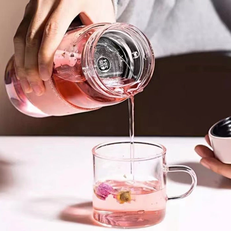 Hand gist rosa Tee aus einer Glasflasche mit integriertem Teesieb in eine klare Teetasse. Im Teesieb sind lose Teekraeuter zu erkennen, und in der Tasse schwimmt eine getrocknete Bluete. Der Hintergrund ist unscharf mit neutralen Farben.