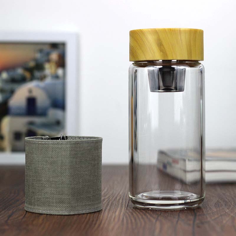 Eine leere, durchsichtige Glas-Trinkflasche mit Bambusdeckel und einem abnehmbaren grauen Stoffband. Neben der Flasche liegt ein integriertes Teesieb, das perfekt in den Flaschenhals passt. Das Bild zeigt die Flasche in einer haeuslichen Umgebung mit einem Bilderrahmen im Hintergrund.