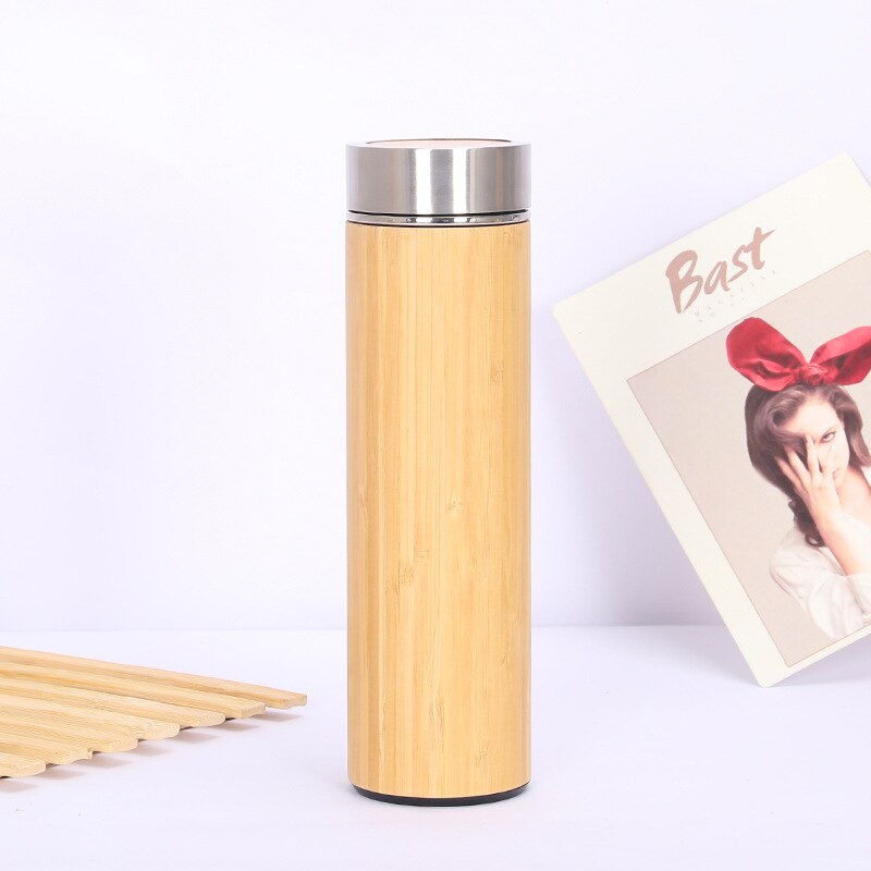 Eine Thermosflasche mit einer Bambushuelle und einem Edelstahldeckel. Im Hintergrund ist eine Postkarte mit einer Person und dem Schriftzug "Bast" zu sehen.
