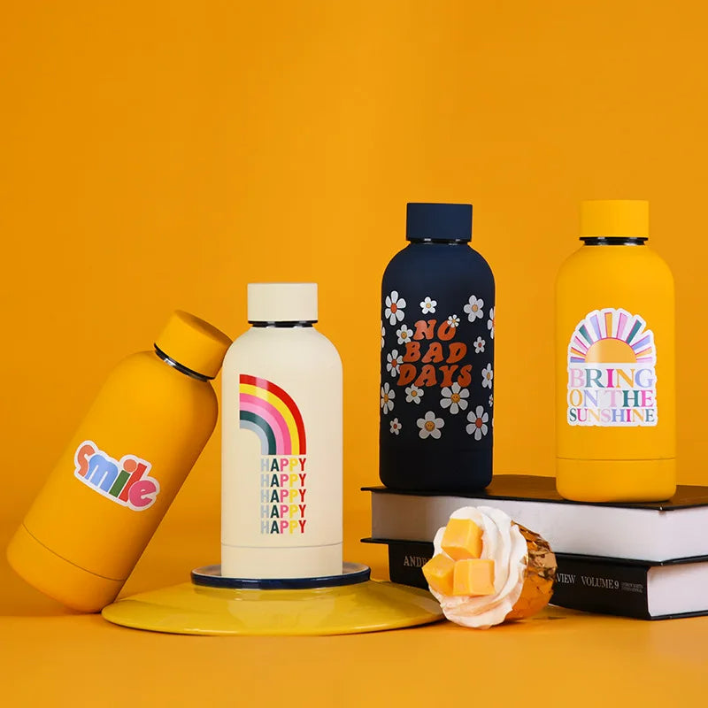 Farbige Thermosflaschen mit unterschiedlichen Designs, darunter Smileys, Schachbrettmuster mit einem Smiley, bunte Blumen auf einem Schachbretthintergrund, "Here Comes The Sun" und ein Regenbogen mit der Aufschrift "Happy", praesentiert auf einem gelben Hintergrund.