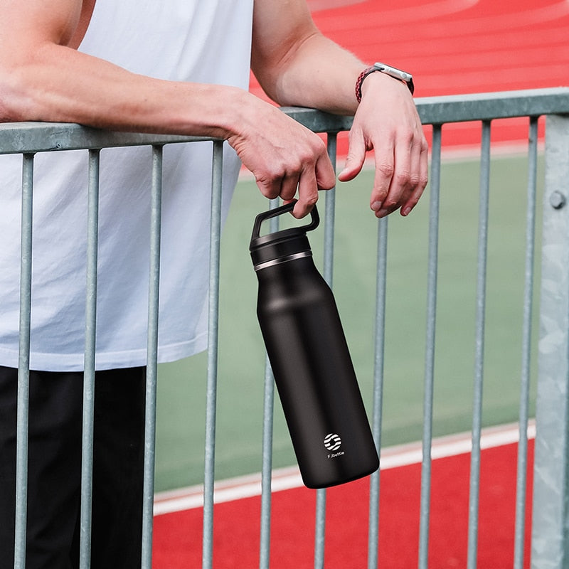 Eine Person, haelt eine schwarze Edelstahl-Thermosflasche an einem Gitterzaun auf einem Sportplatz.