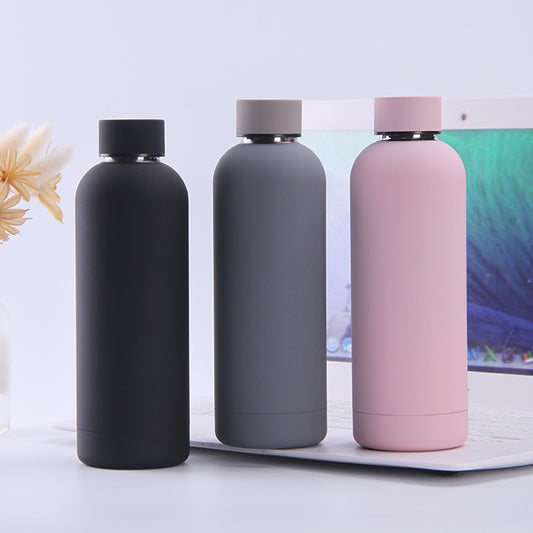 Drei matte Thermosflaschen in Schwarz, Grau und Rosa, platziert neben einem Laptop.