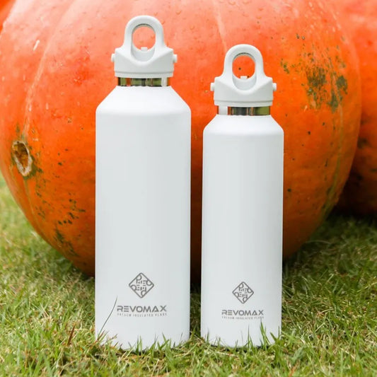 Zwei weiße Edelstahl-Thermosflaschen der Marke Revomax, vor einem großen Kuerbis auf Gras
